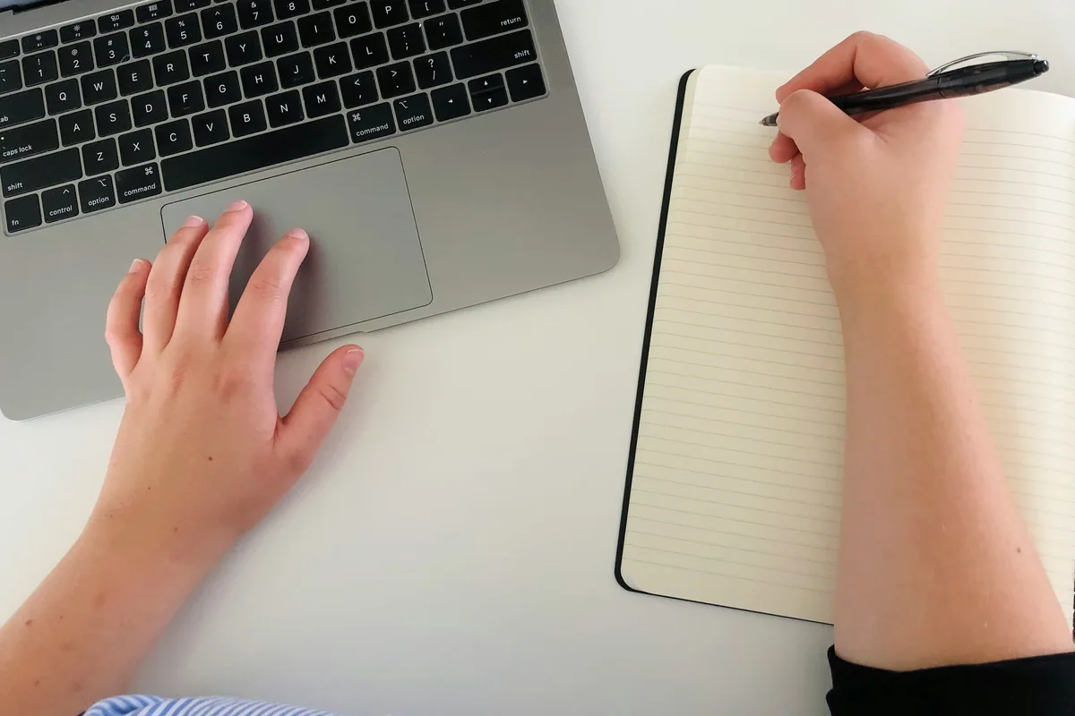 Mãos junto ao teclado de um notebook e a um caderno aberto, com uma caneta.