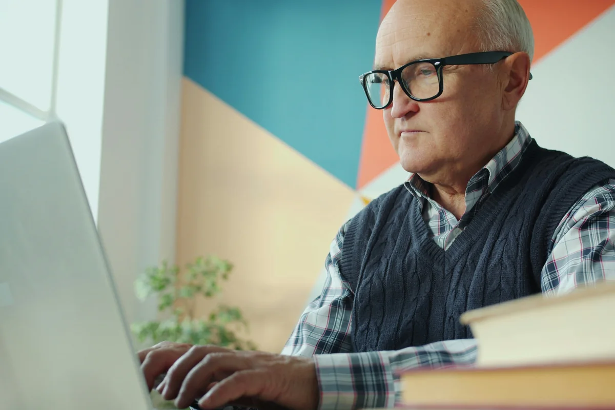 Homem de cabelos grisalhos e óculos sentado diante de um notebook, com livros sobre a mesa.