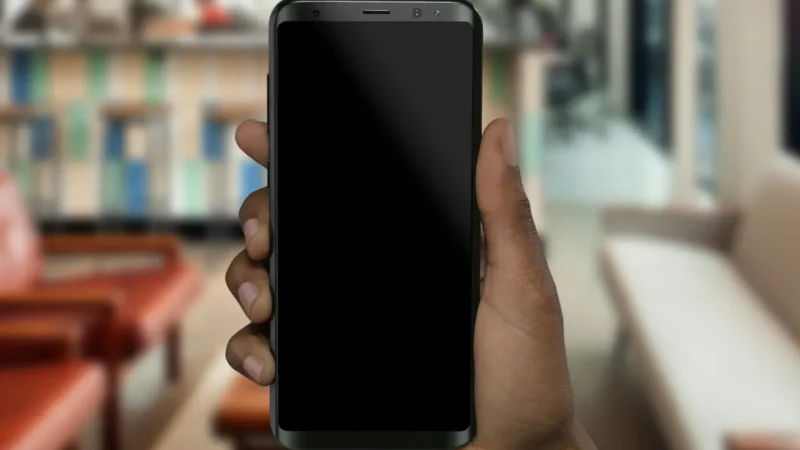 Mão segura um celular com a tela apagada em um espaço de trabalho desfocado.