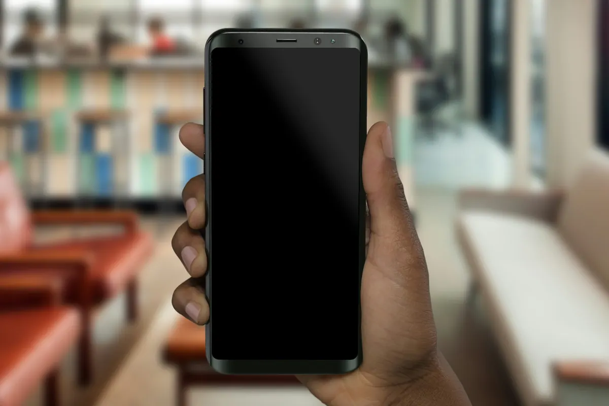 Mão segura um celular com a tela apagada em um espaço de trabalho desfocado.